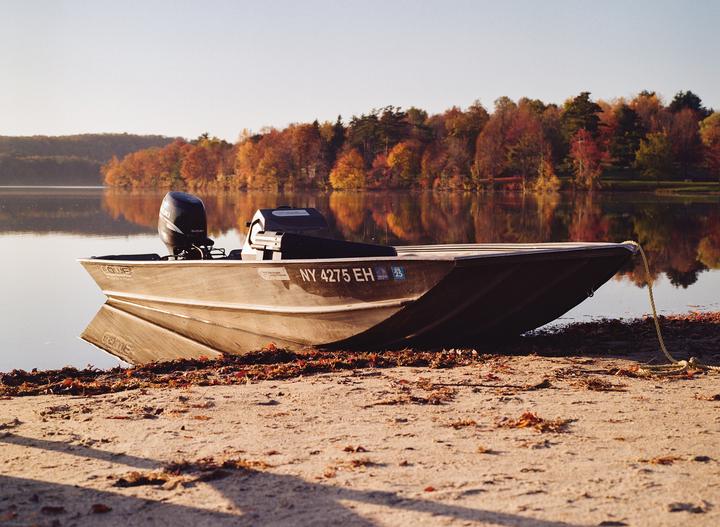 A boat is pulled onto a sandy shore next to a lake with autumn trees reflecting in the water.