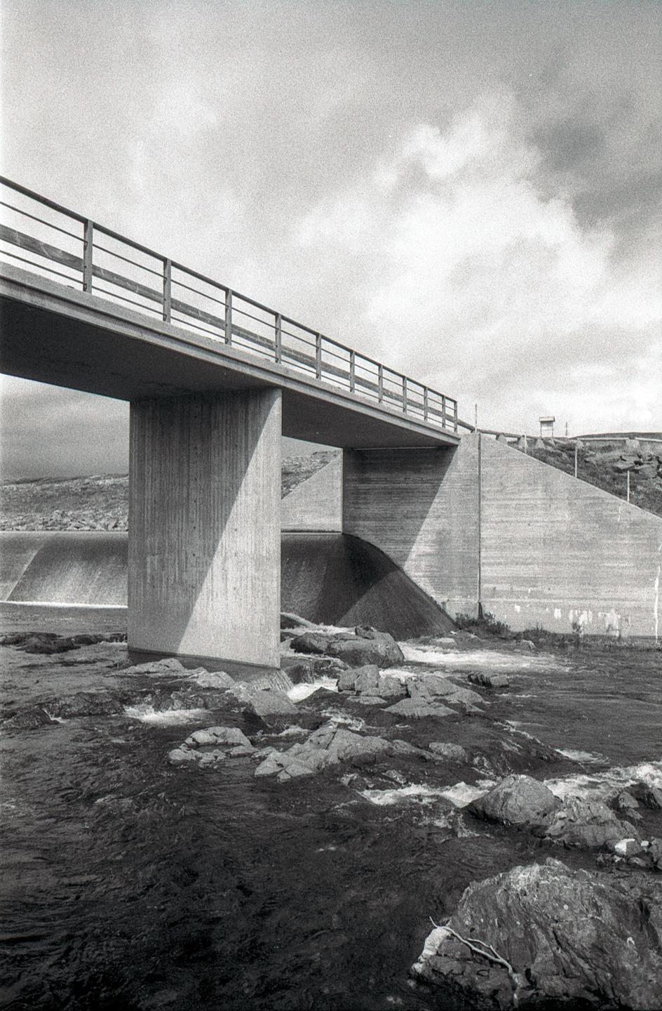 A concrete bridge with a metal railing spans over a river with rocks and a small waterfall.