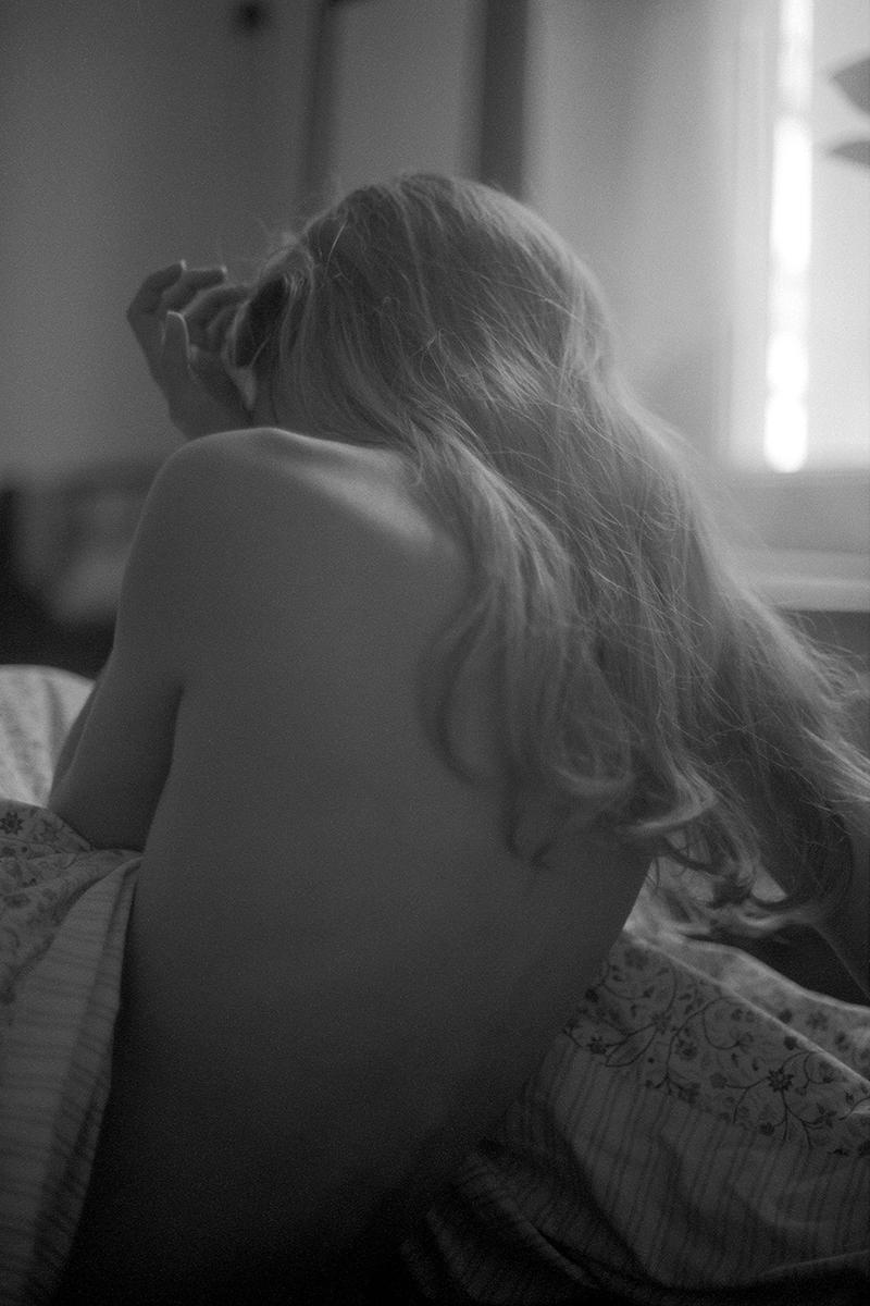 A woman with long hair sits on a bed with her back to the viewer, looking towards a window.