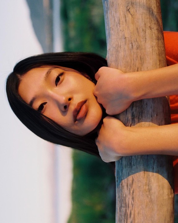 A woman with dark hair leans her head against a wooden post, looking directly at the viewer.