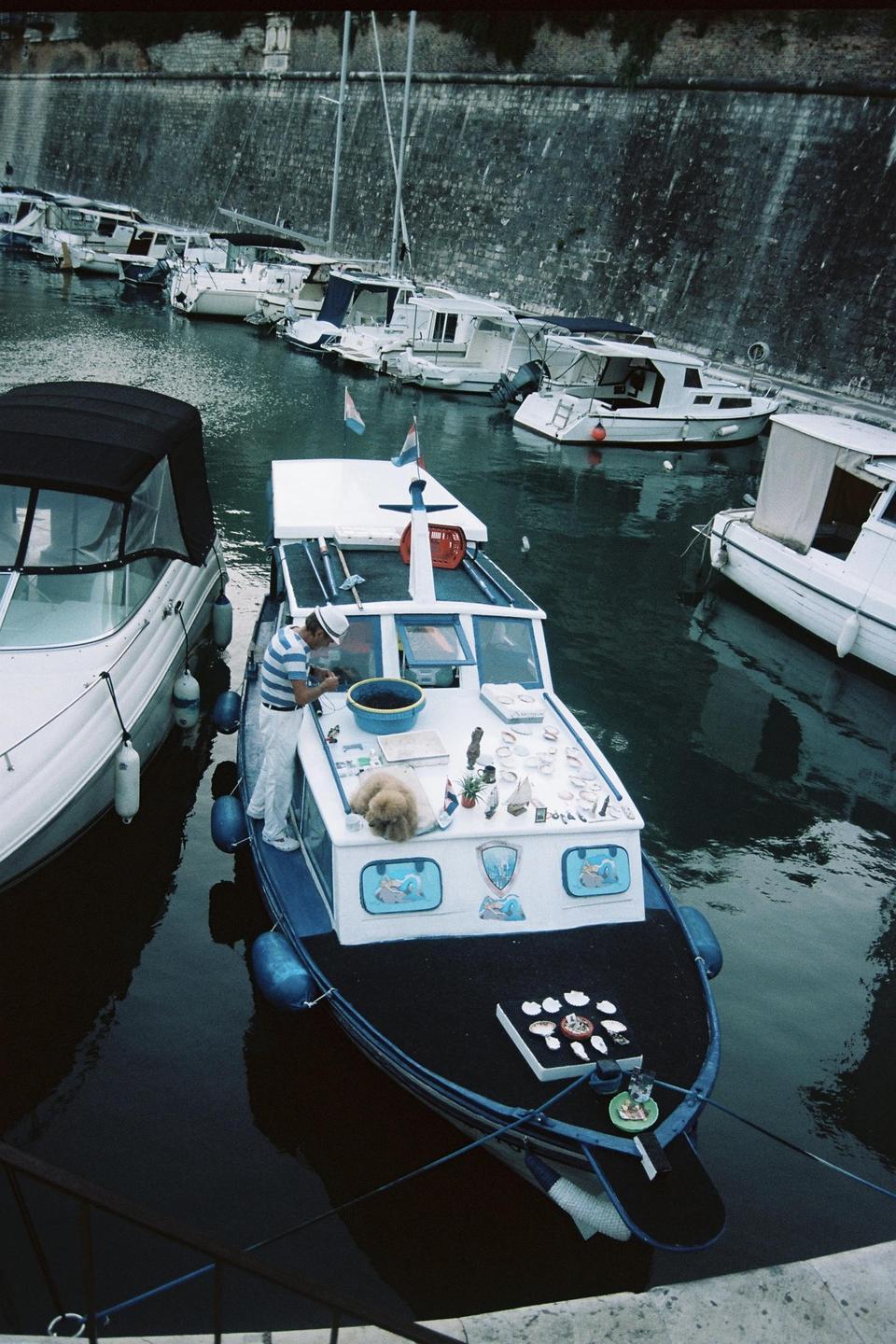 A man stands on a boat selling items from its deck, with a dog sleeping nearby and other boats docked in the background.