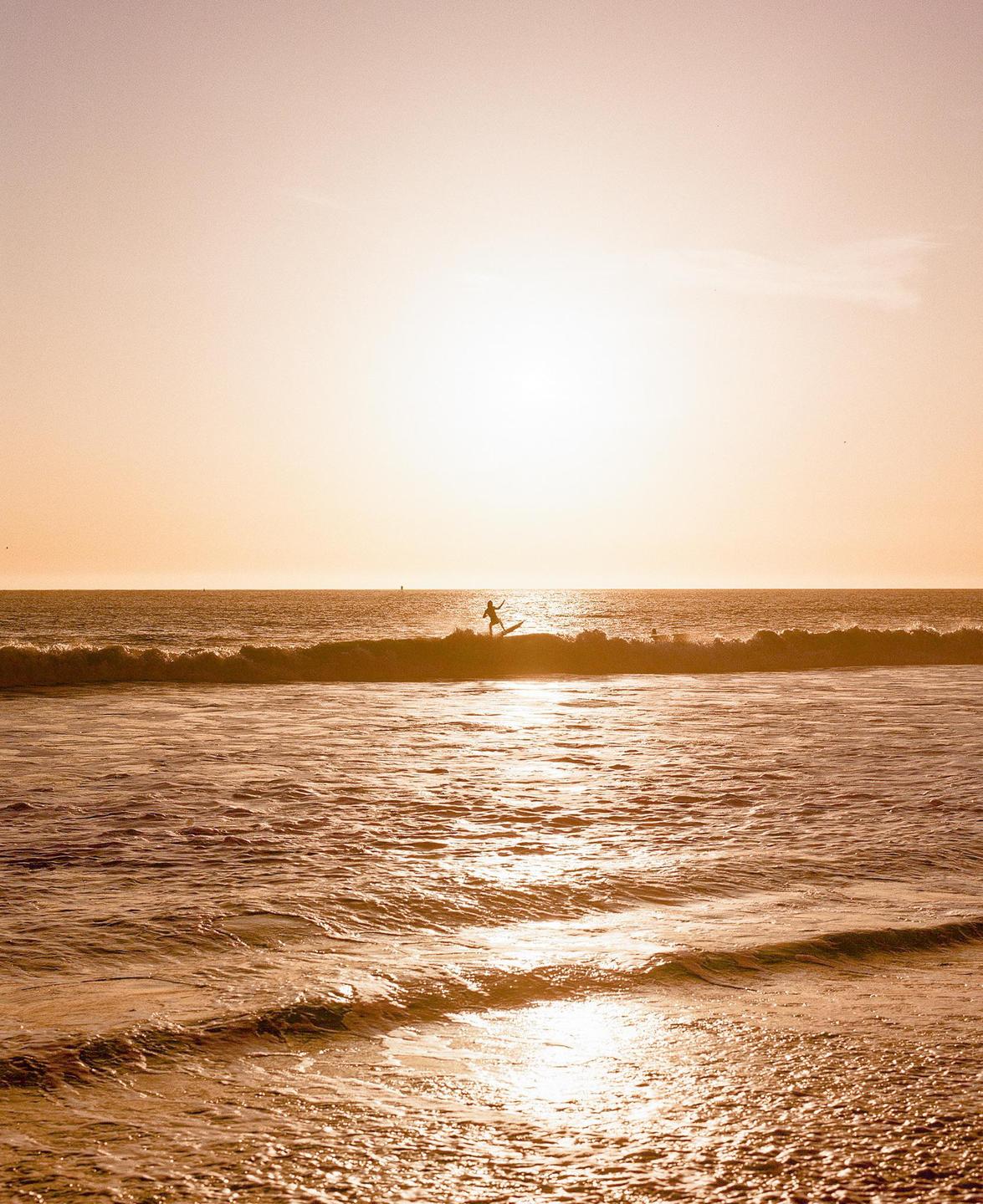 A surfer rides a wave at sunset, silhouetted against the bright sky.