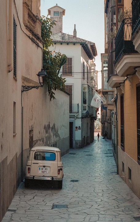 A small white car is parked on a narrow cobblestone street lined with tall buildings.
