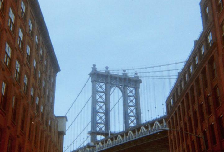 A large bridge is visible between two tall brick buildings under a light blue sky.