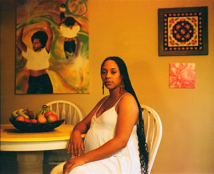 A pregnant woman with long braids sits at a table with a bowl of fruit, with paintings on the wall behind her.