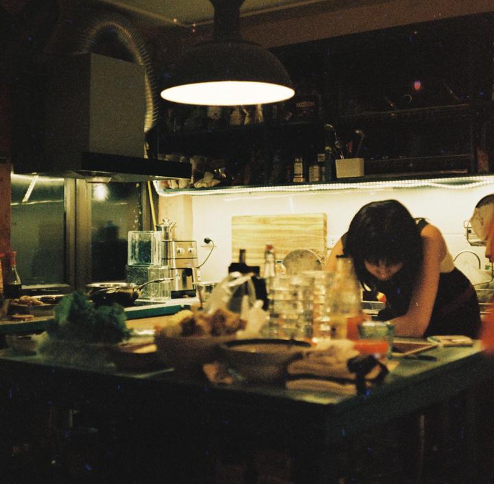 A woman leans over a kitchen counter filled with dishes and food under a bright overhead light.