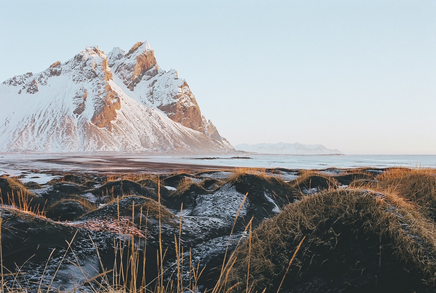 A snow-covered mountain range rises behind a coastal landscape with grassy dunes and a calm ocean under a clear sky.