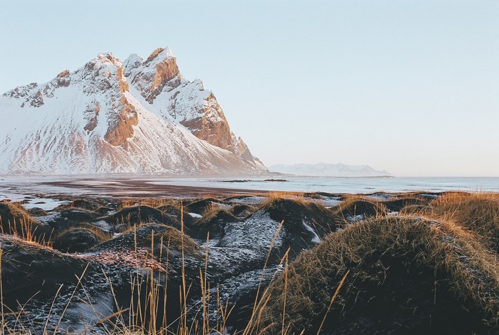 A snow-covered mountain range rises behind a coastal landscape with grassy dunes and a calm ocean under a clear sky.