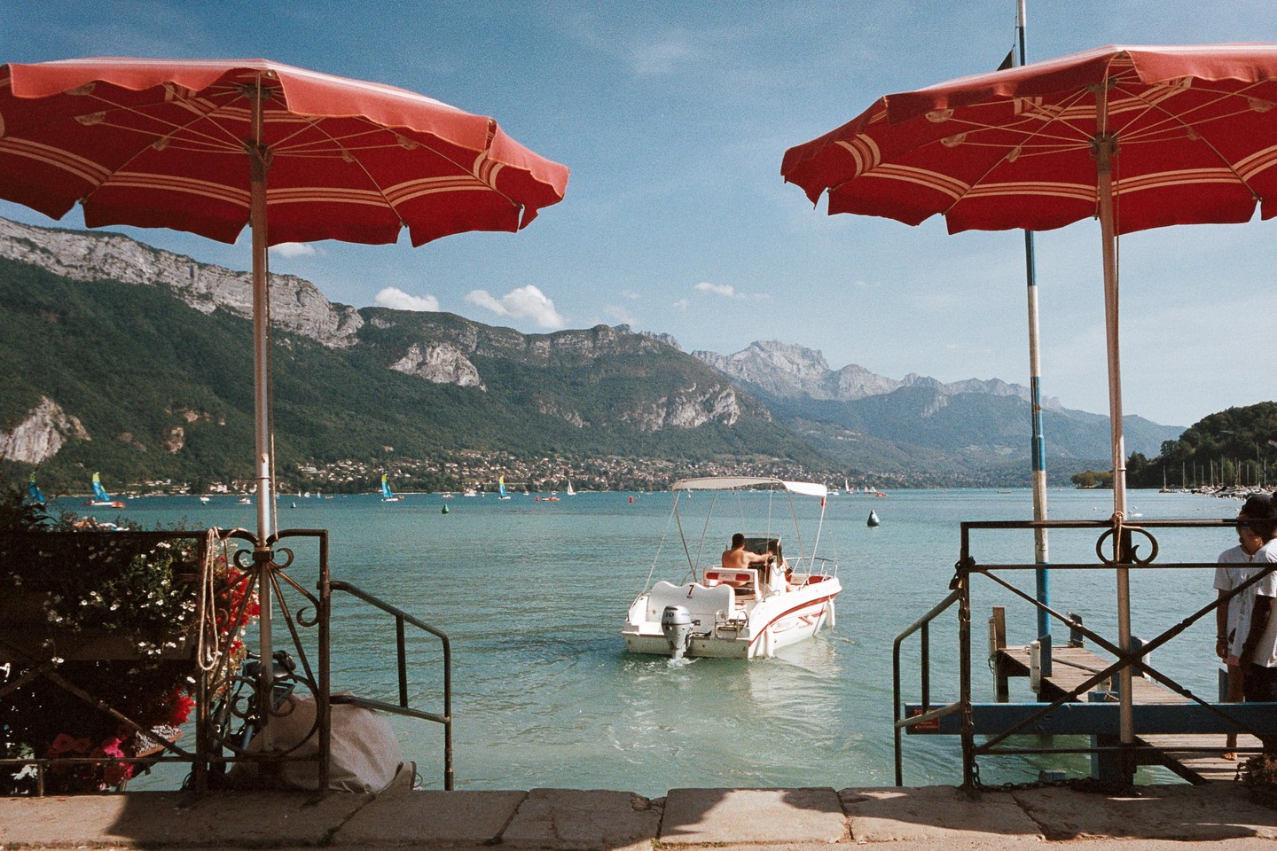 Two red umbrellas frame a lake with mountains in the background and a boat with a man on it.