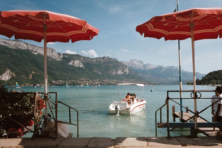Two red umbrellas frame a lake with mountains in the background and a boat with a man on it.