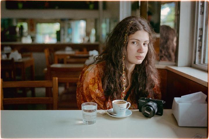 A young woman with long, curly hair sits at a table in a bar with a cup of coffee and a glass of water.