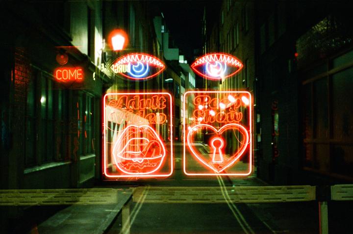 Neon signs for an adult video store and peep show illuminate a dark street at night.
