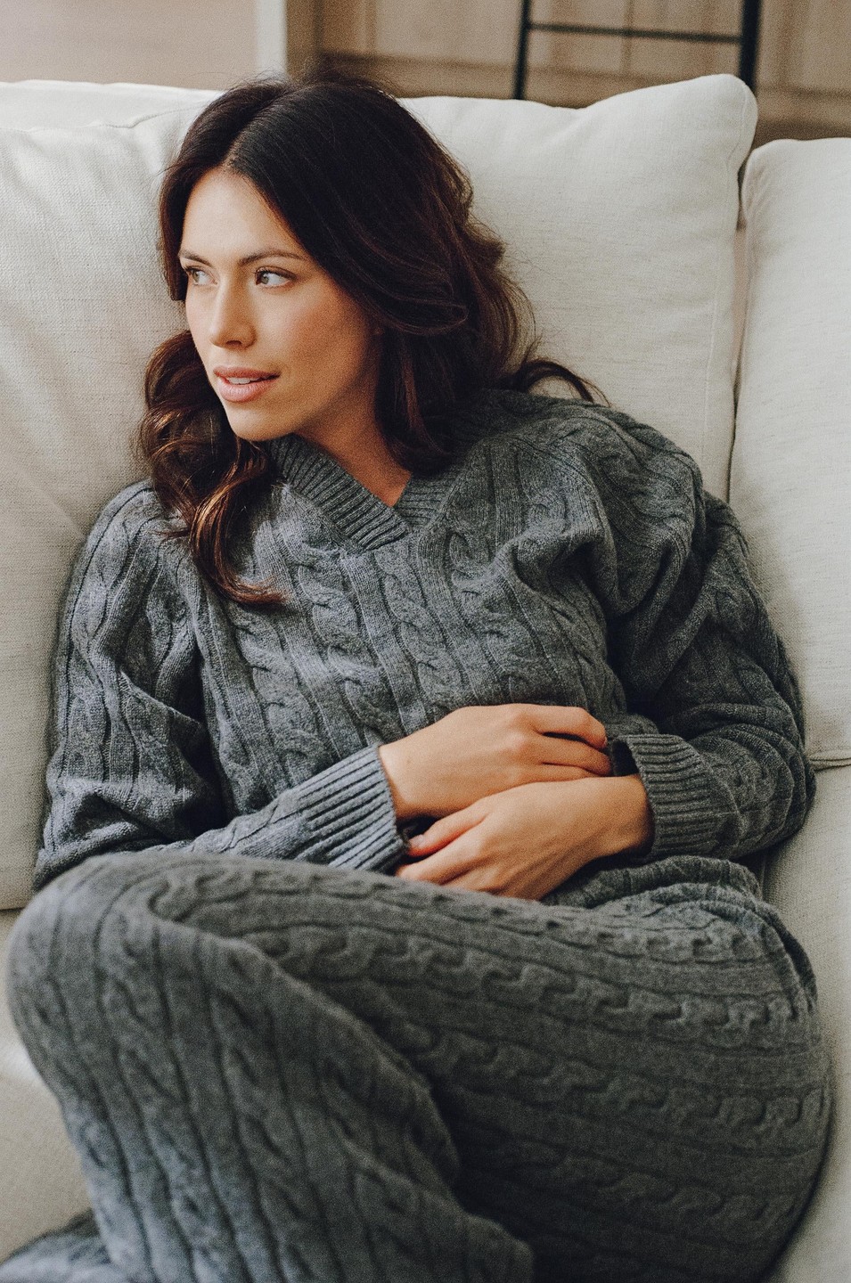 A woman in a gray cable knit sweater and pants is reclining on a sofa.