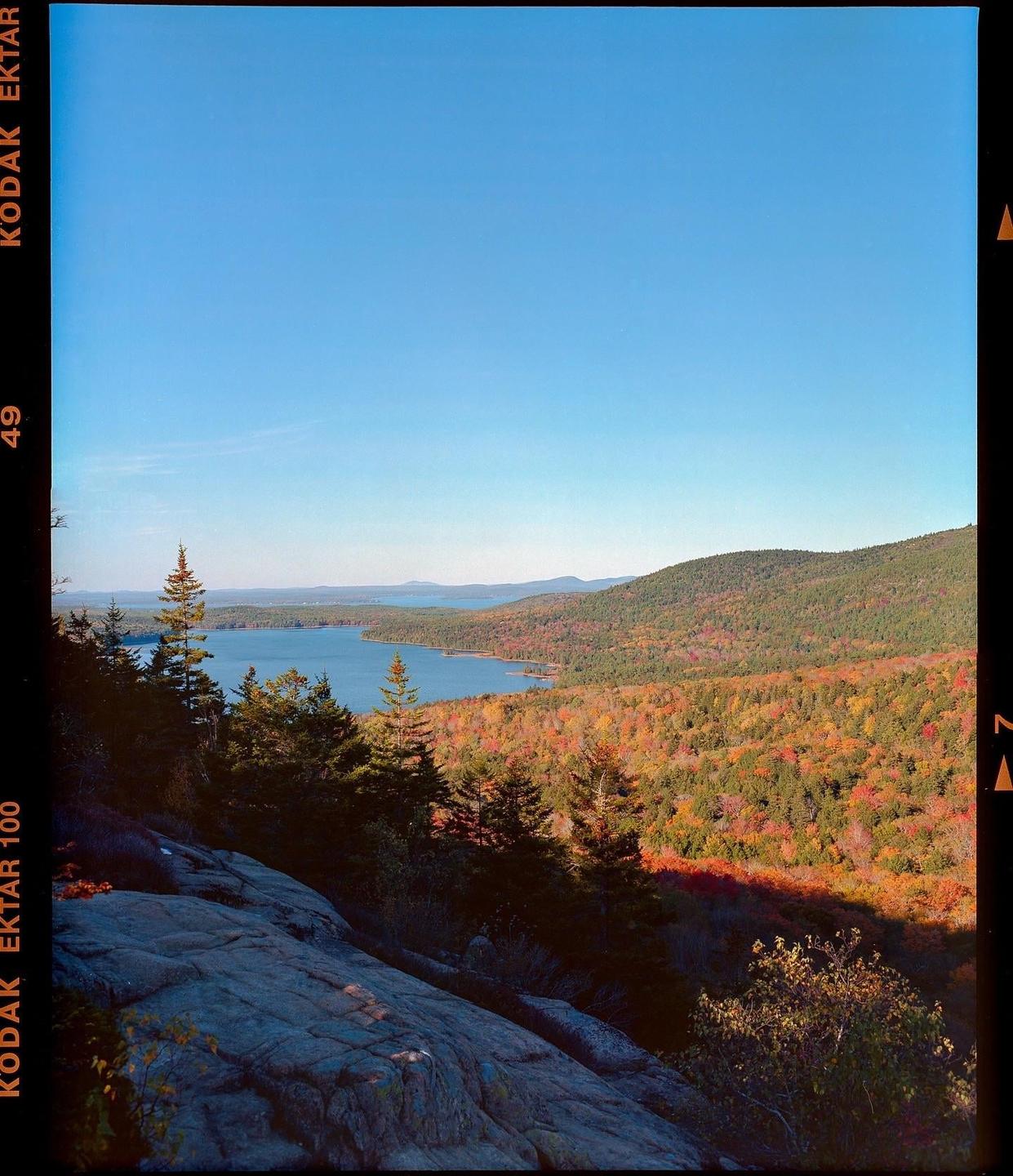 A scenic landscape shows a body of water surrounded by hills covered in autumn foliage under a clear blue sky.