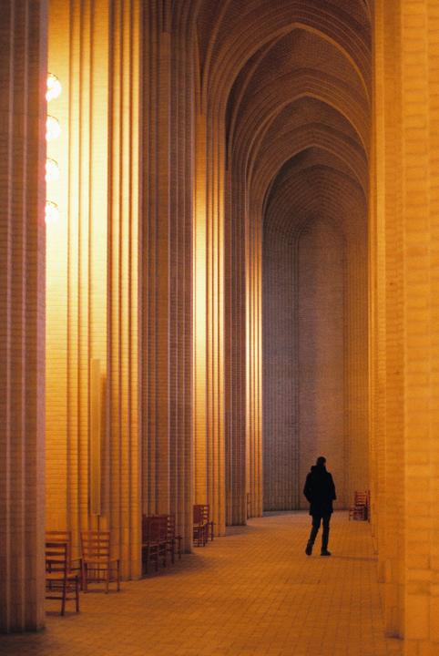 A person walks down a long, arched hallway with tall, narrow windows on one side and chairs lined up against the wall.