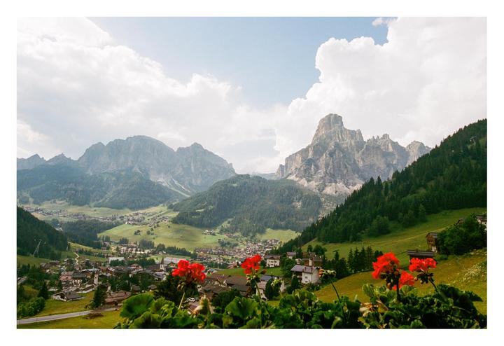 Red geraniums frame a view of a mountain village nestled in a valley with towering peaks under a cloudy sky.