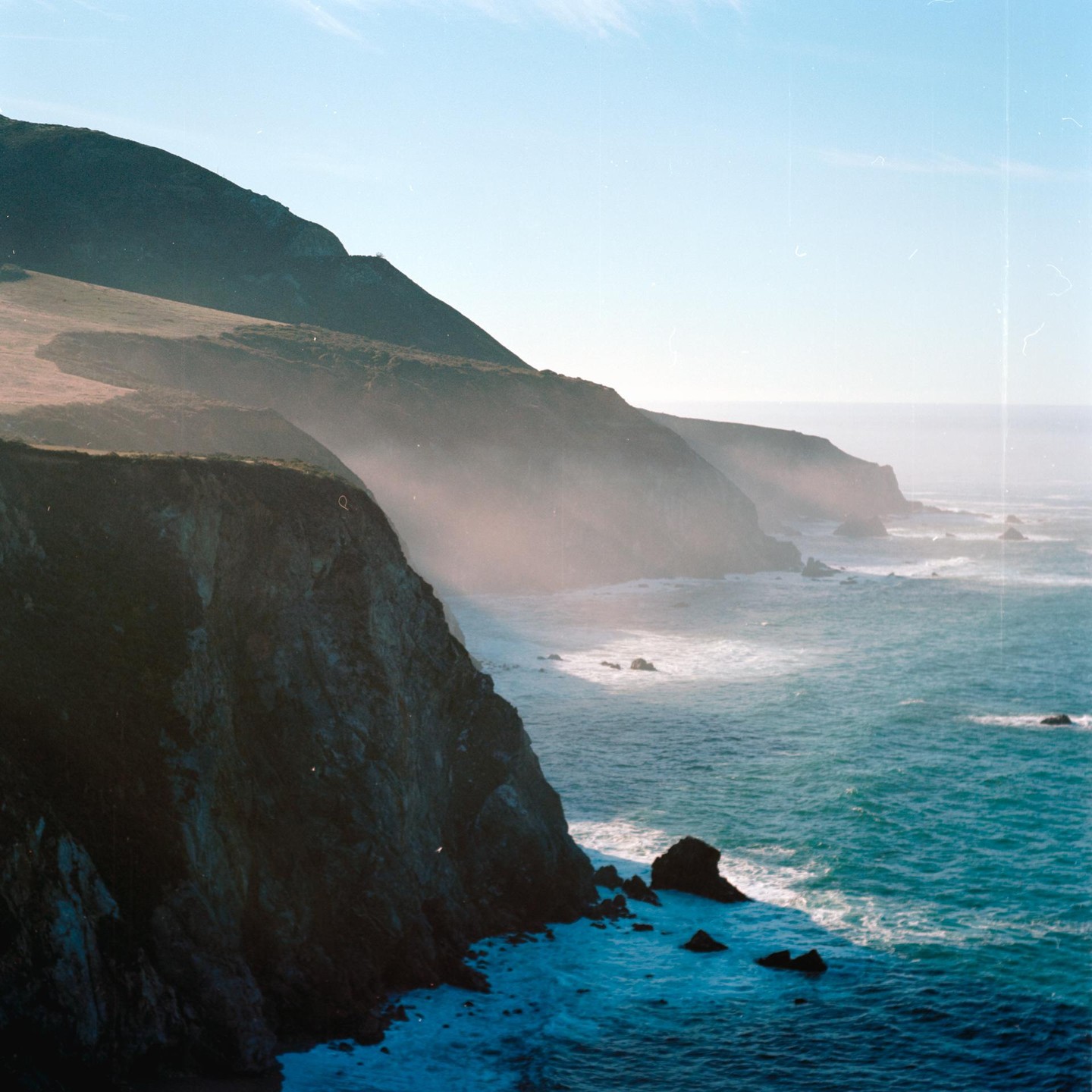The morning sun illuminates the misty coastline of Big Sur, California, with waves crashing against the rocky shore.