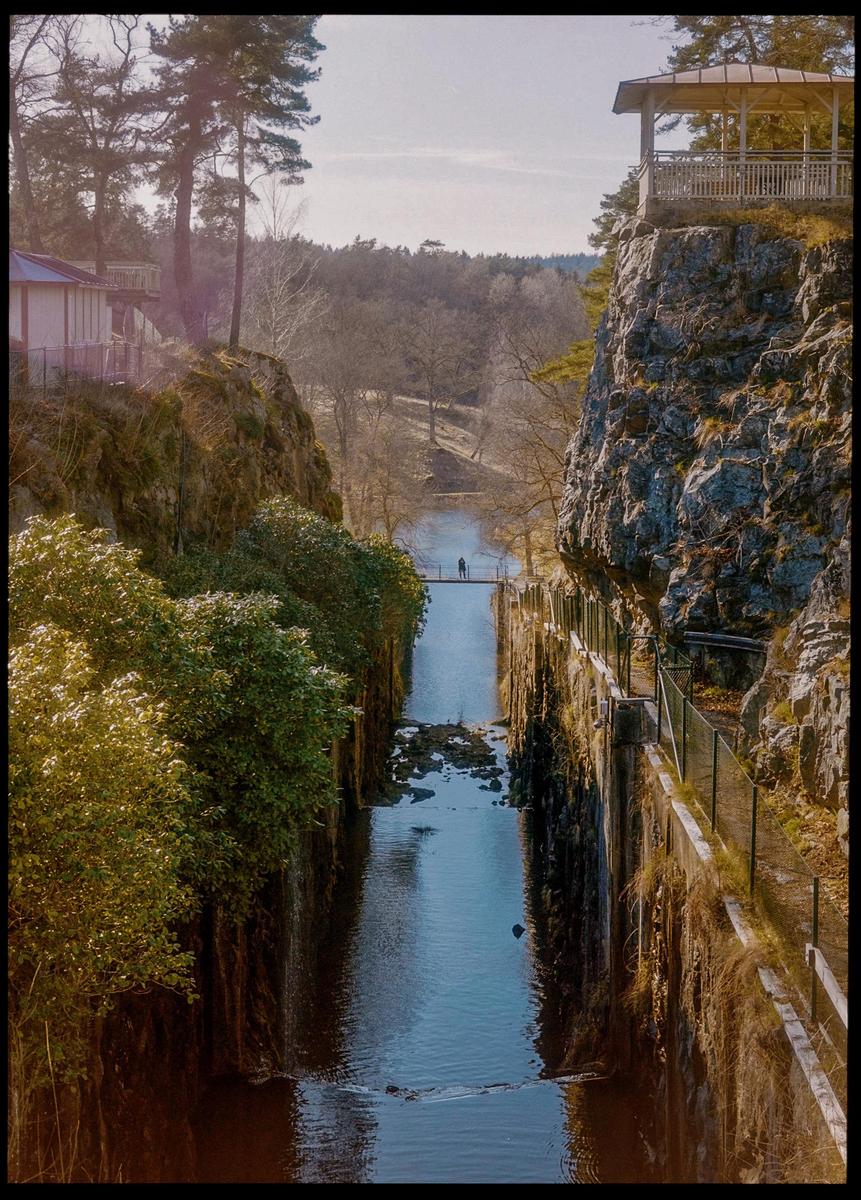 A narrow river flows between two steep, rocky banks, with a small bridge and a person visible in the distance.