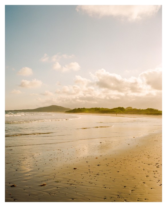 A person walks along a sandy beach with waves crashing on the shore and mountains in the distance under a cloudy sky.