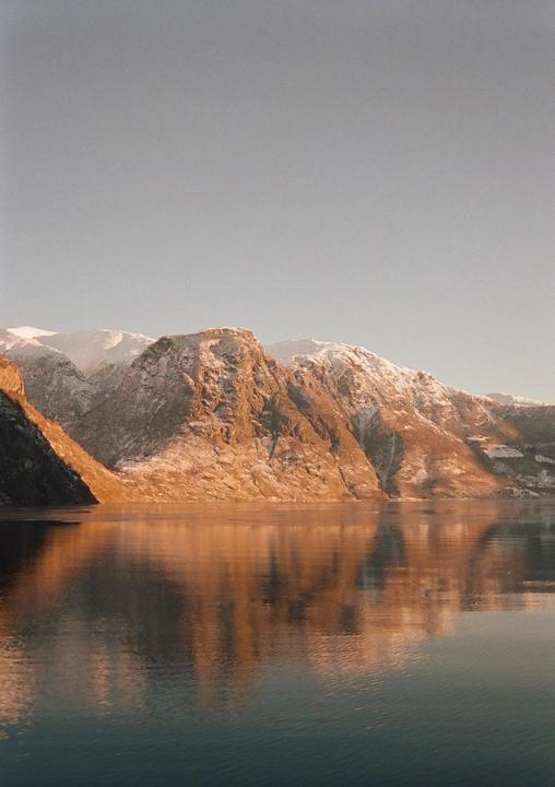 Snow-capped mountains are reflected in the still water of a fjord during golden hour.