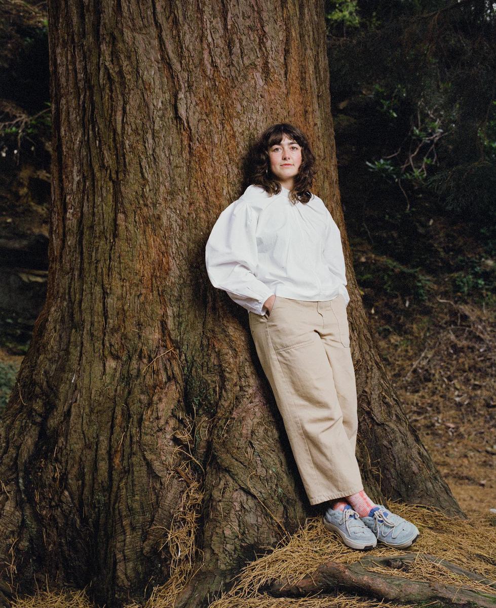 A woman with dark, curly hair leans against a large redwood tree.