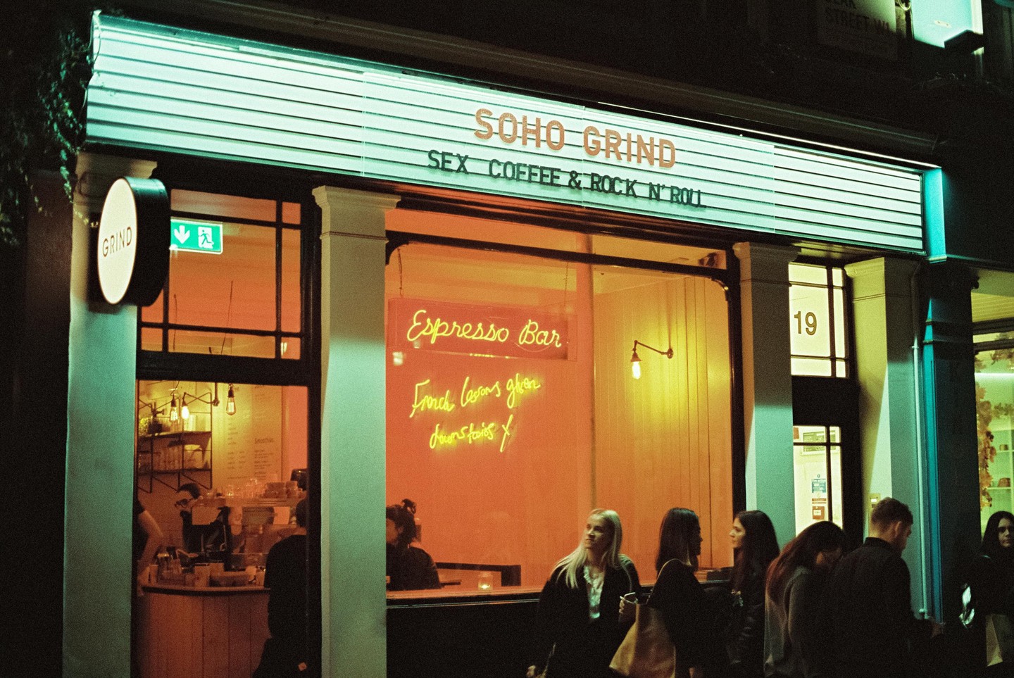 A group of people stand outside a cafe with a neon sign that reads "Espresso Bar" and "Soho Grind" at night.