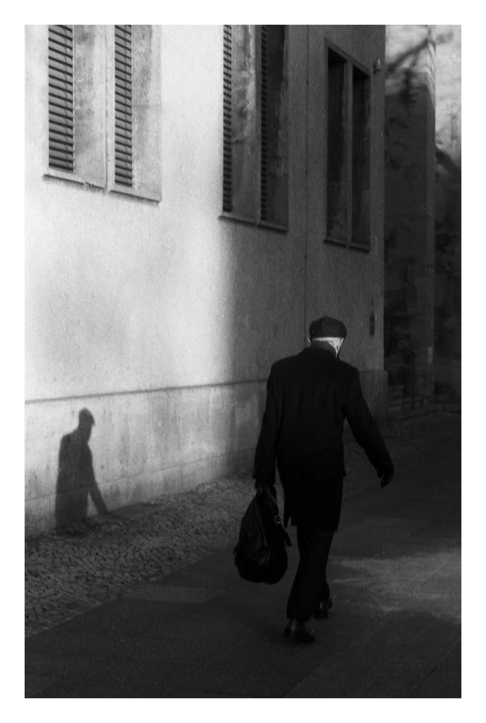 A man walks away from the camera on a street next to a building with his shadow cast on the wall.