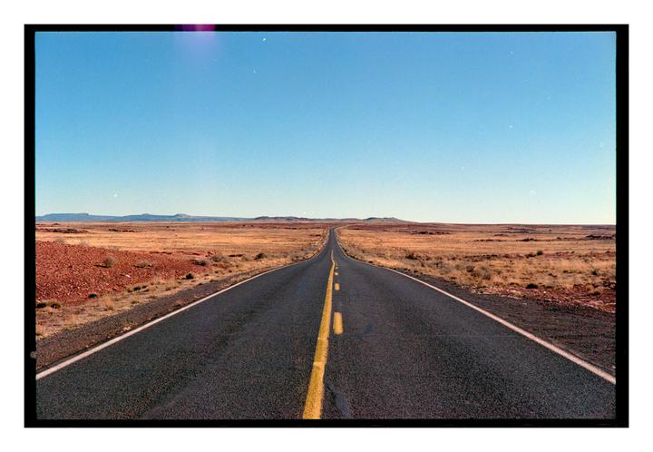 A long, straight road stretches into the distance through a dry, open landscape under a clear sky.
