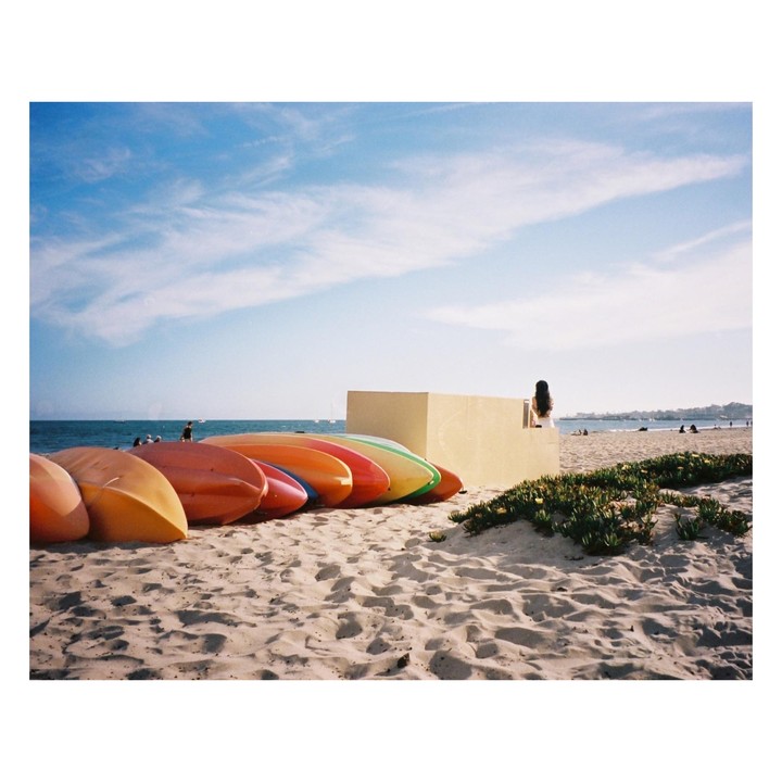 A woman sits on a low wall on a sandy beach next to colorful kayaks.