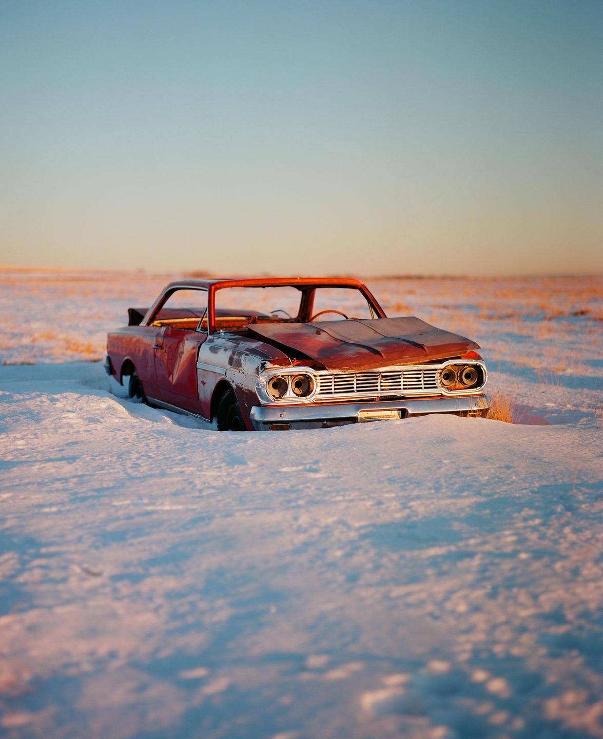 A rusty red car is abandoned in a snowy field under a clear sky.