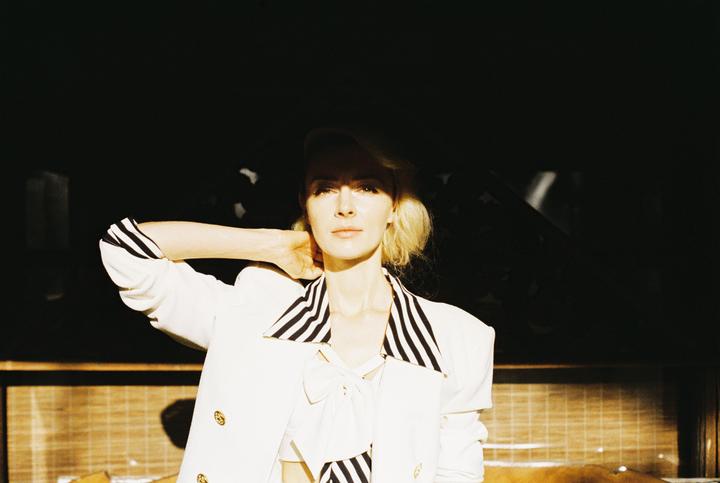 A woman in a white jacket with striped accents poses with her hand behind her head, illuminated by a strong beam of light.