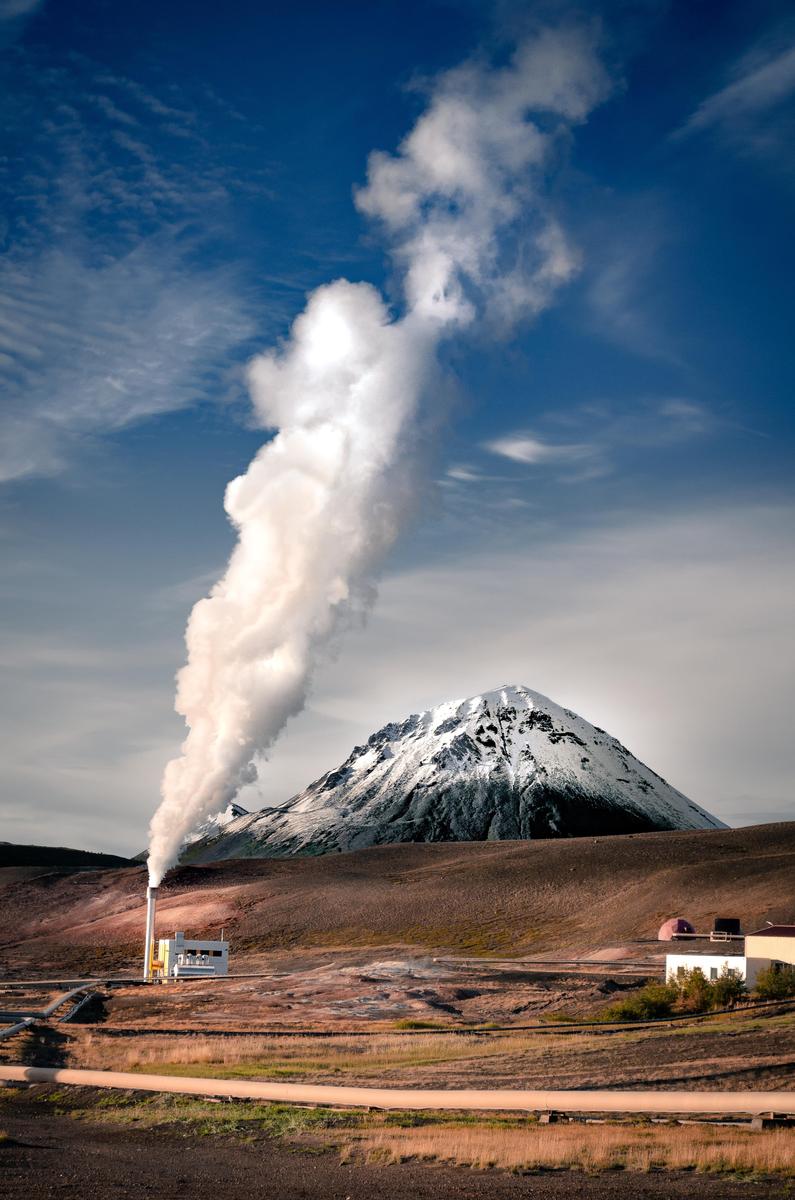 A geothermal power plant emits a large plume of steam next to a snow-capped mountain under a blue sky.