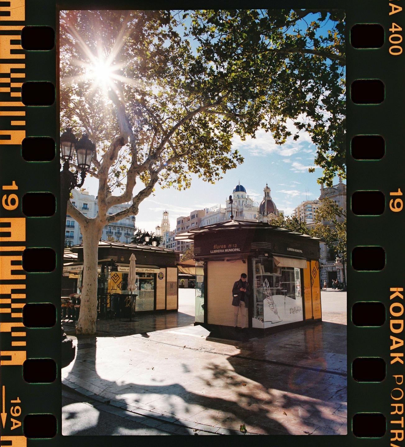 A man stands outside a small shop with a tree and buildings in the background.