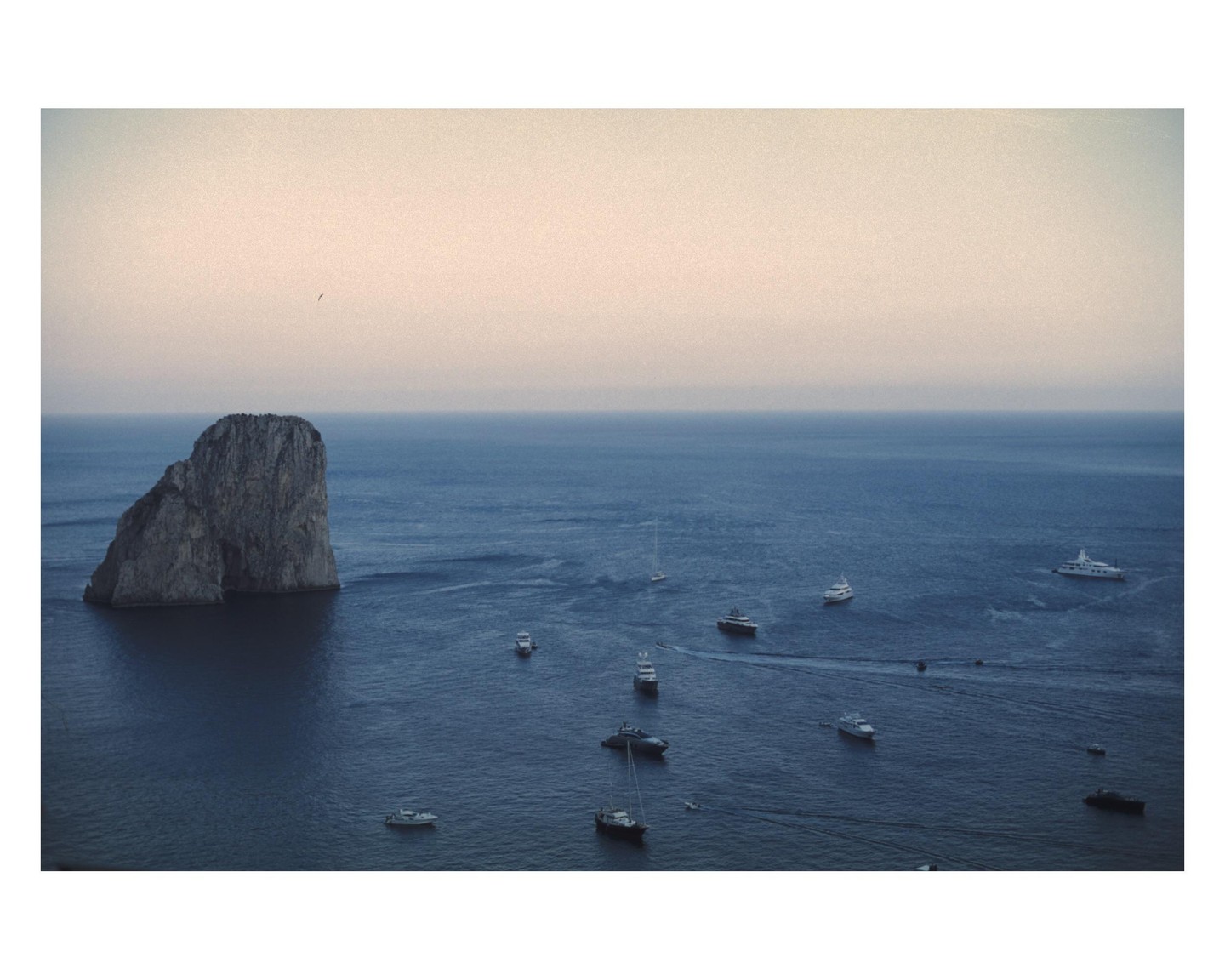 A large rock formation rises from the sea, surrounded by numerous boats under a hazy sky.