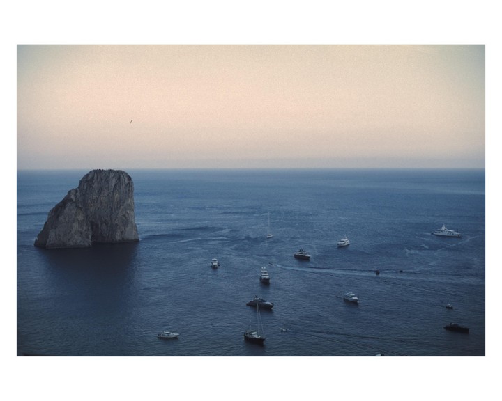 A large rock formation rises from the sea, surrounded by numerous boats under a hazy sky.