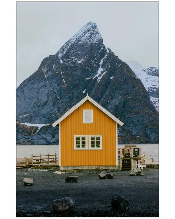 A bright yellow house with white trim stands in front of a large, snow-capped mountain and a body of water.