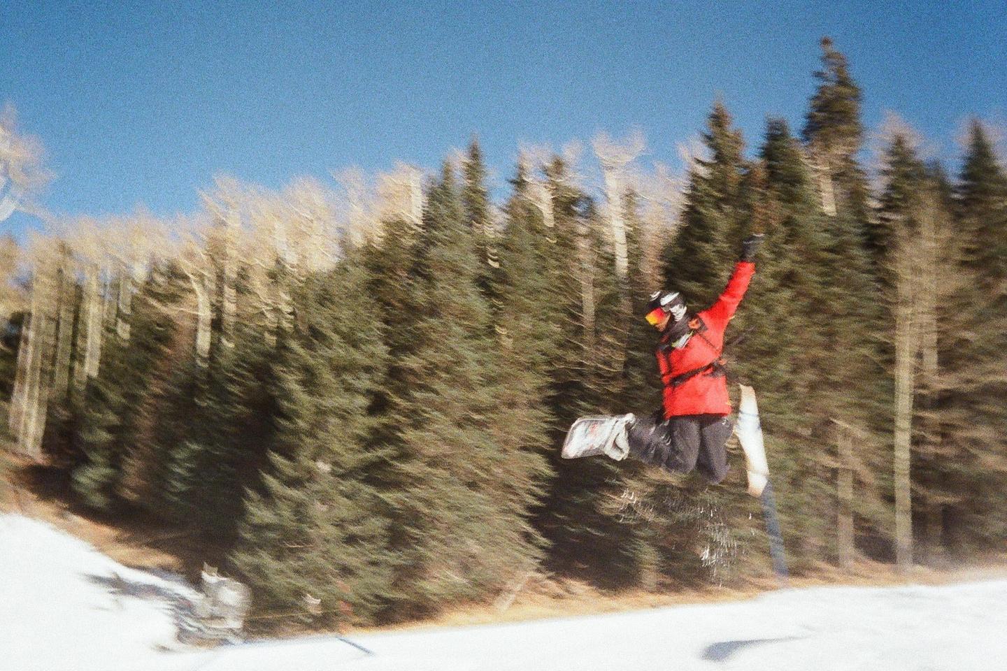 A snowboarder in a red jacket is captured mid-air against a backdrop of pine trees and a clear blue sky.