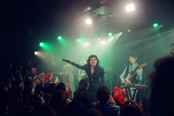 A woman sings into a microphone on stage, surrounded by band members and an audience.