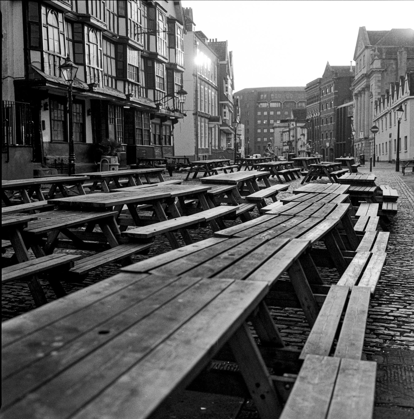 Many empty picnic tables are arranged on a cobblestone street in front of buildings with the sun shining brightly.