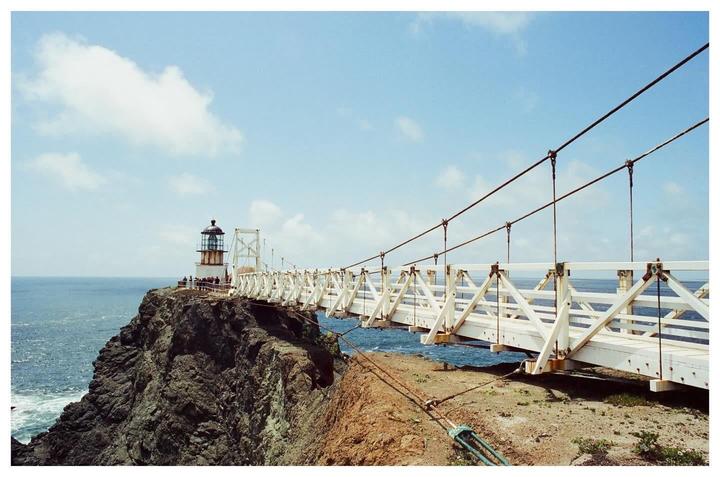 A white suspension bridge leads to a lighthouse on a rocky cliff overlooking the ocean under a blue sky with clouds.