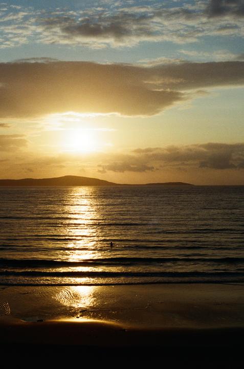 A person is swimming in the ocean with the sun reflecting on the water and hills in the distance.