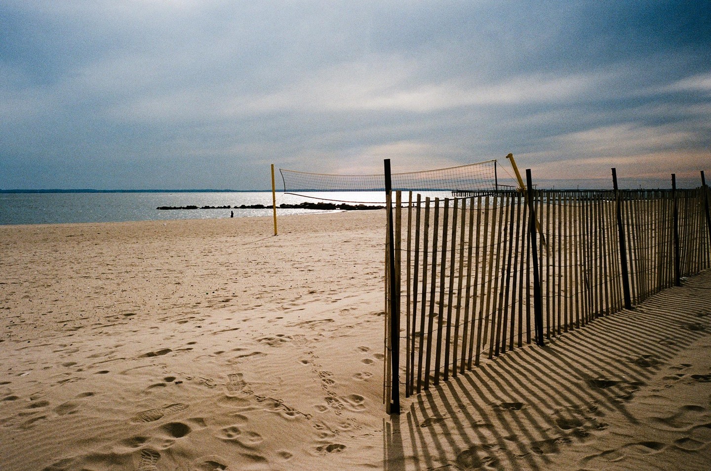 A beach volleyball net stands on a sandy beach next to a wooden fence with long shadows stretching across the sand.
