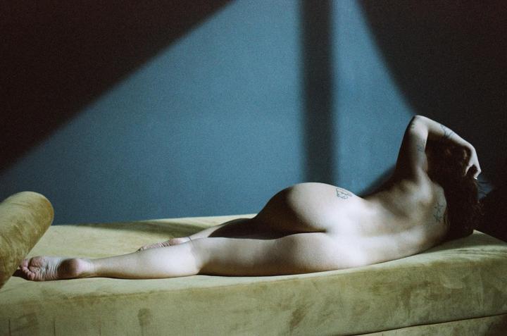 A woman lies nude on a yellow couch with a blue wall behind her, illuminated by a diagonal beam of light.
