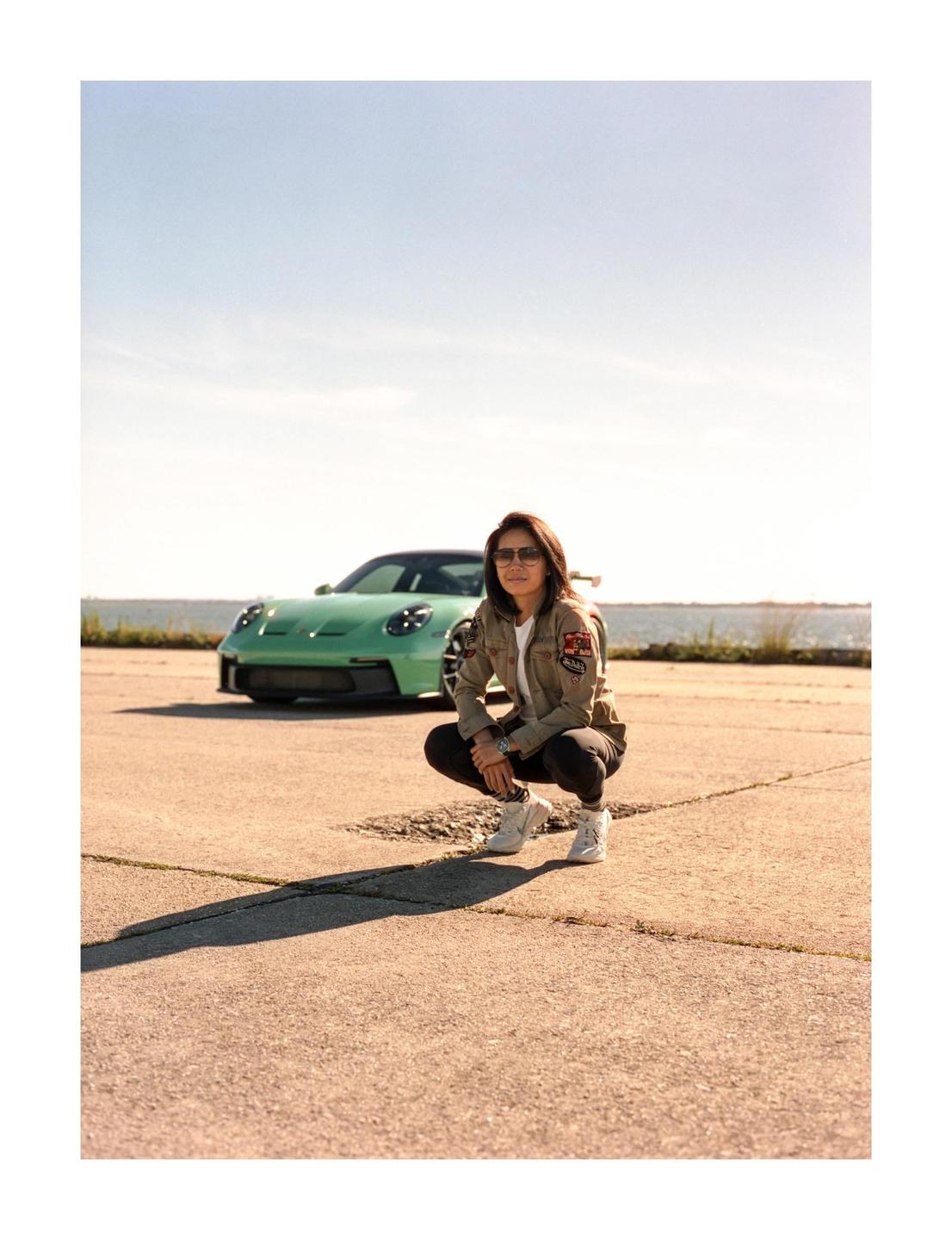 A woman squats in front of a green sports car with a body of water in the background.