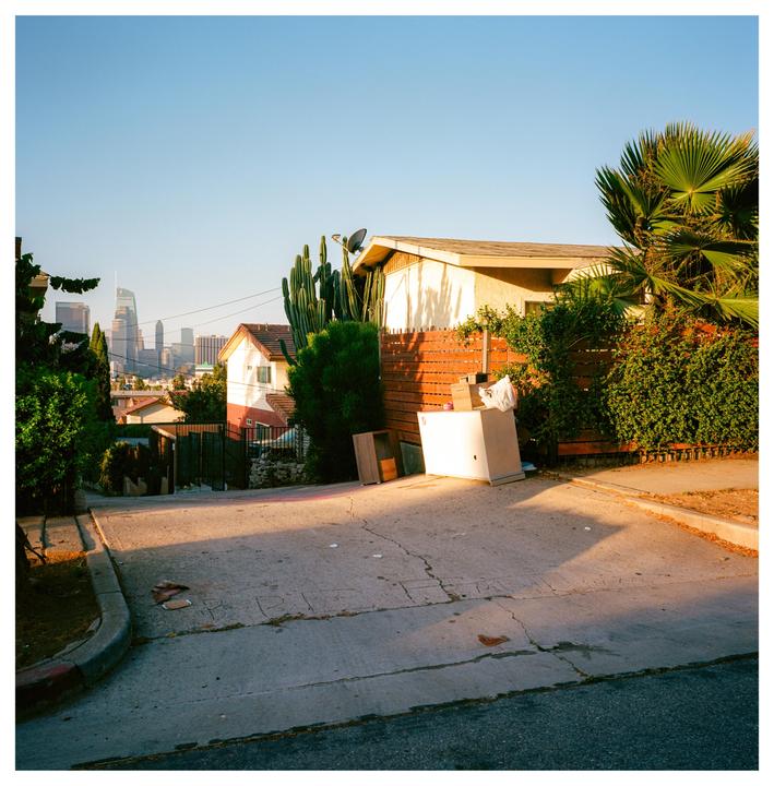 A residential street with houses and lush greenery leads down to a distant city skyline under a clear sky.
