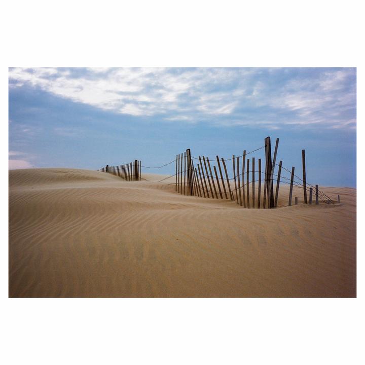 A wooden fence with wire runs across a sandy dune under a cloudy sky.