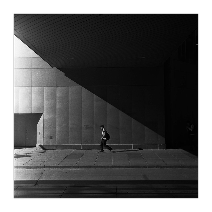 A man walks along a sunlit sidewalk next to a building with a large shadow cutting across its facade.
