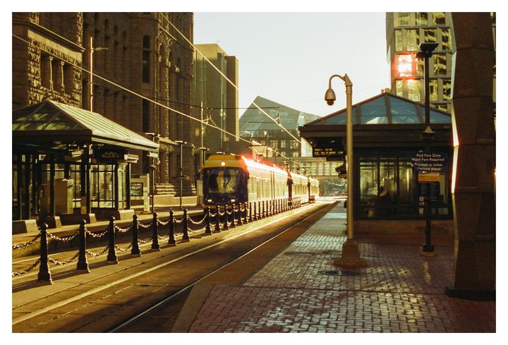 A train approaches a station platform on a sunny morning, with buildings and overhead wires visible in the background.
