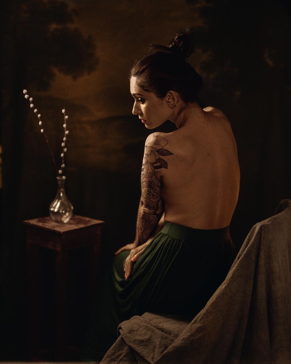 A woman with a tattoo on her arm sits with her back to the viewer, looking over her shoulder at a vase with branches on a small table.