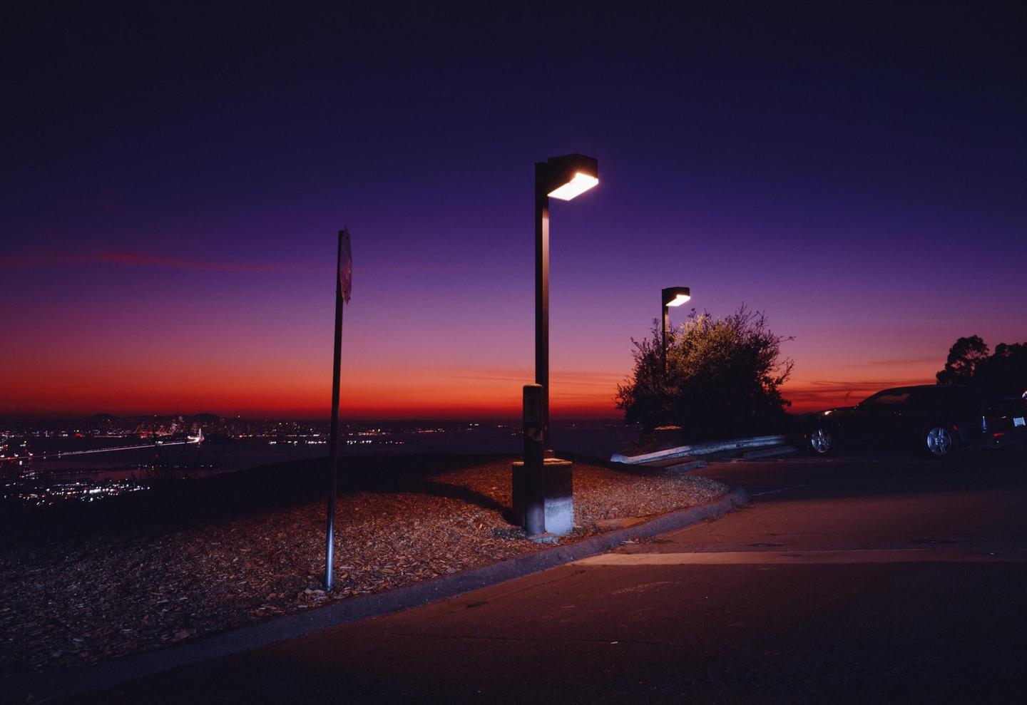 A street light illuminates a parking area overlooking a city at sunset.
